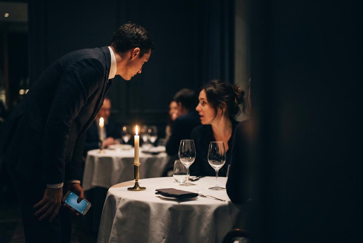 A maître d’ leaning in to hear a guest at a candle-lit table, a phone in hand.